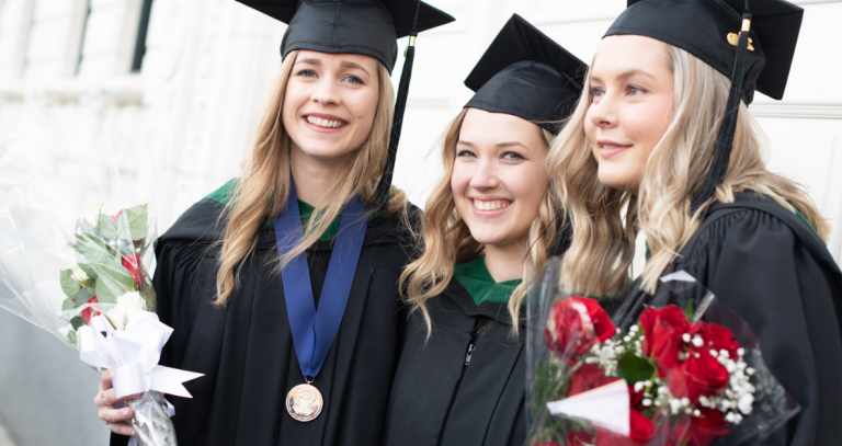 Three graduates smile while holding flowers in their caps and gowns. One is wearing a convocation medal.